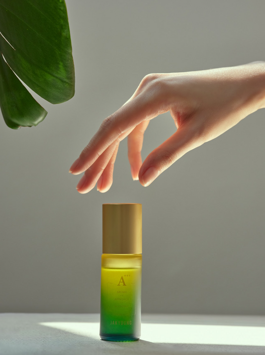 A hand hovers above a small cosmetic bottle with a gold cap and a green-to-yellow gradient body, set against a soft gray background with a large green leaf in the upper left.