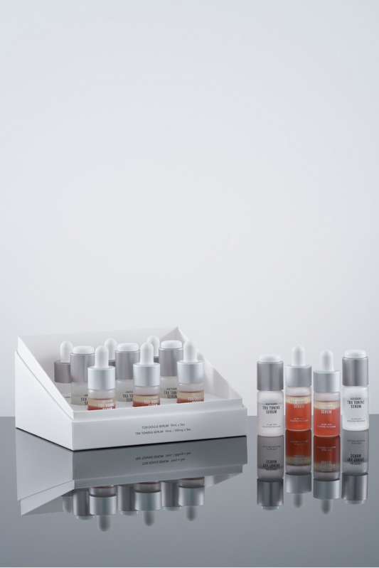 A skincare set featuring a white display tray and six small dropper bottles with silver caps and amber liquid, arranged on a reflective dark surface against a light gray background.