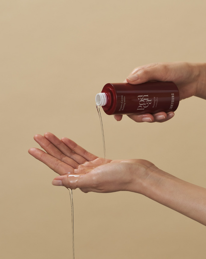 A person's hands holding a dark red bottle and pouring a clear gel-like product into their palm against a solid beige background.