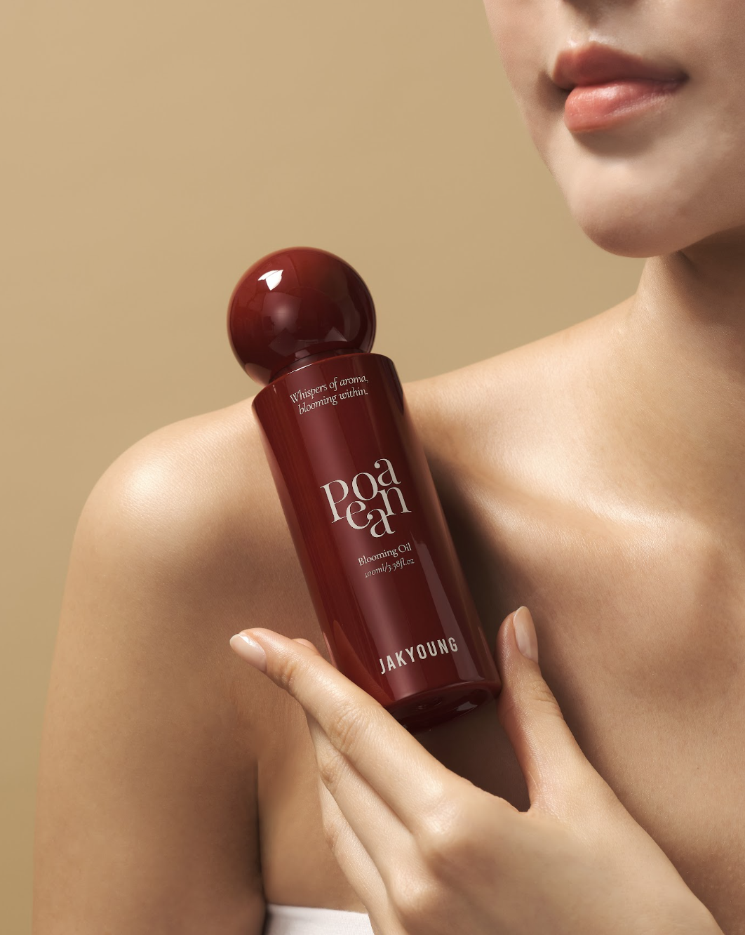 A close-up of a model holding a dark red bottle of Janyoung Poedam Blushing Oil against a solid beige background.
