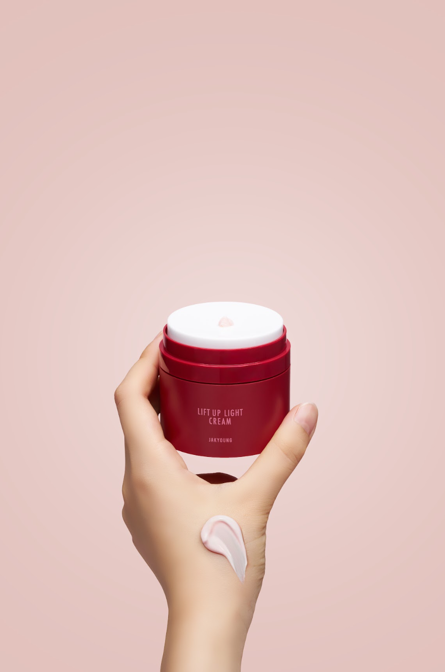 A hand holding a red cosmetic jar labeled 'LIFT UP LIGHT CREAM' against a soft pink background, with a dollop of white cream on the back of the hand.