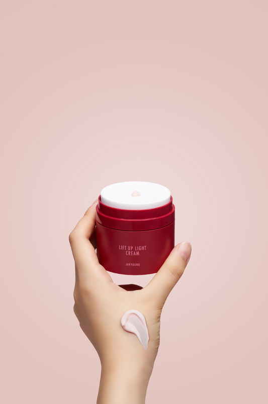 A hand holding a red cosmetic jar labeled 'LIFT UP LIGHT CREAM' against a soft pink background, with a dollop of white cream on the back of the hand.