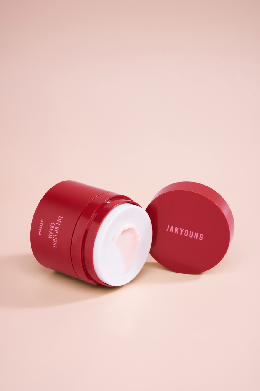 A red cosmetic jar of JAKYOUNG Cream with its lid off, showing the white cream inside, set against a solid pale pink background.