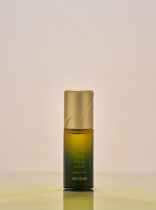 A bottle of Jakyung Energy Oil with a gold cap and a dark green-to-amber gradient bottle, centered on a light beige background with a soft reflection below.