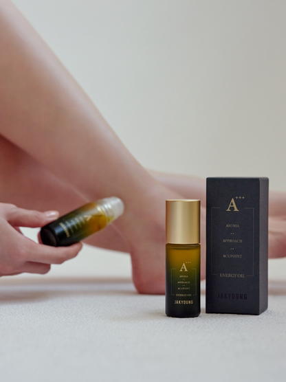 A small amber glass perfume or essential oil bottle with a gold cap stands next to its dark charcoal box, while a person's hands and legs hold a matching bottle in the background against a soft beige backdrop.