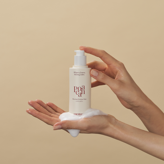 A person's hands holding a bottle with white foam dripping from the bottom into the palm. The background is a solid, warm beige color.