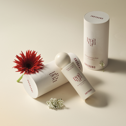Three white JAKYOUNG skincare containers arranged on a warm cream background with a red flower and small white blossoms as styling props. 